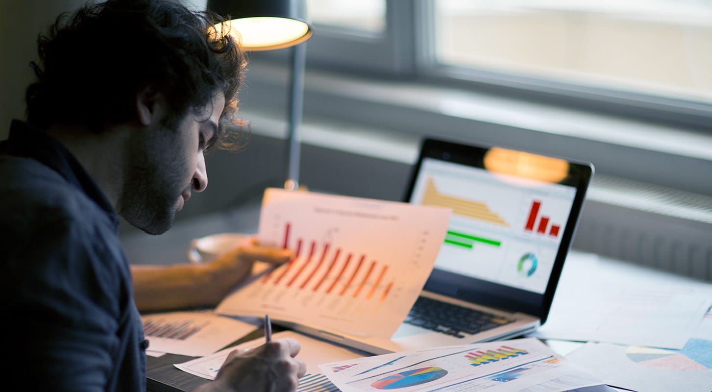 A man sits at a desk with an open laptop, analyzing papers with bar graphs and notes around him.